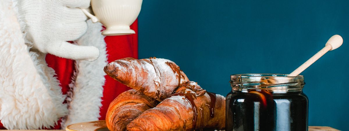 Father Christmas holds a cup of tea next to a plate of croissants and jar full of jam.