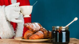 Father Christmas holds a cup of tea next to a plate of croissants and jar full of jam.