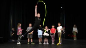 A young performer jumps whilst twirling a ribbon as other young people look on a photograph from Empire Youth Theatre.